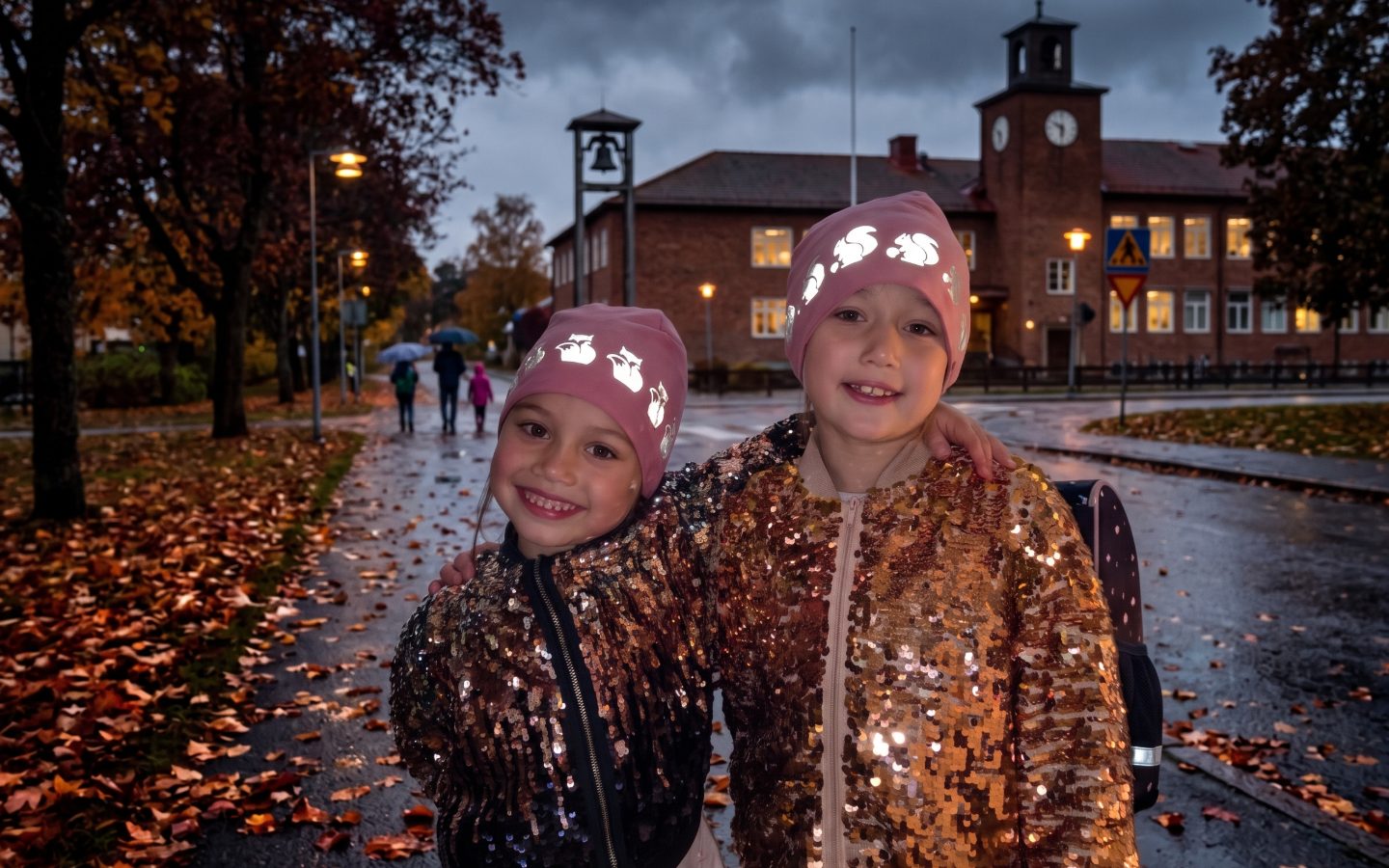 To piger med lyserøde reflekshuer foran skolen på en mørk efterårsdag for optimal trafiksikkerhed.