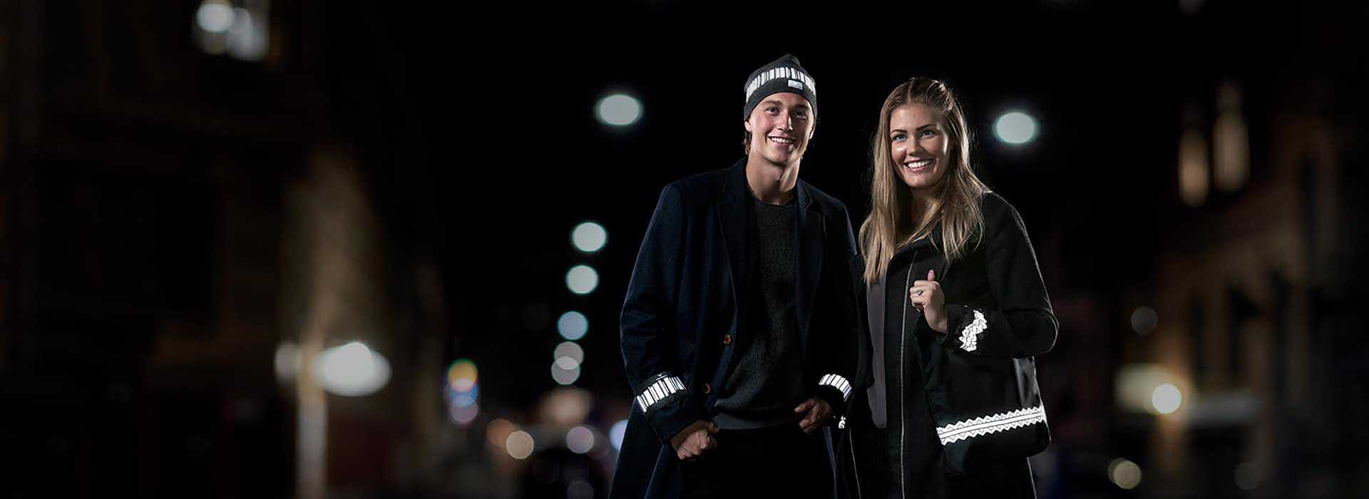 Man och kvinna. Han bär mössa med reflexmönstret norrsken. Hon håller i Reflexväska med mönstret Fjälltopp. Båda bär ärmreflex